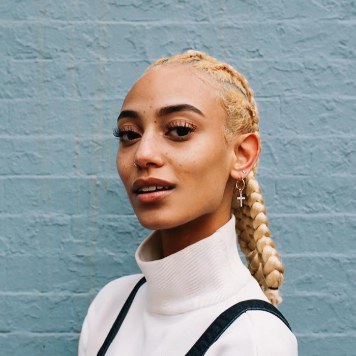 Vintage toned portrait of a young mixed race Latina woman from New York. She is sporting striking blond braids, leaning against the pastel toned brick wall.